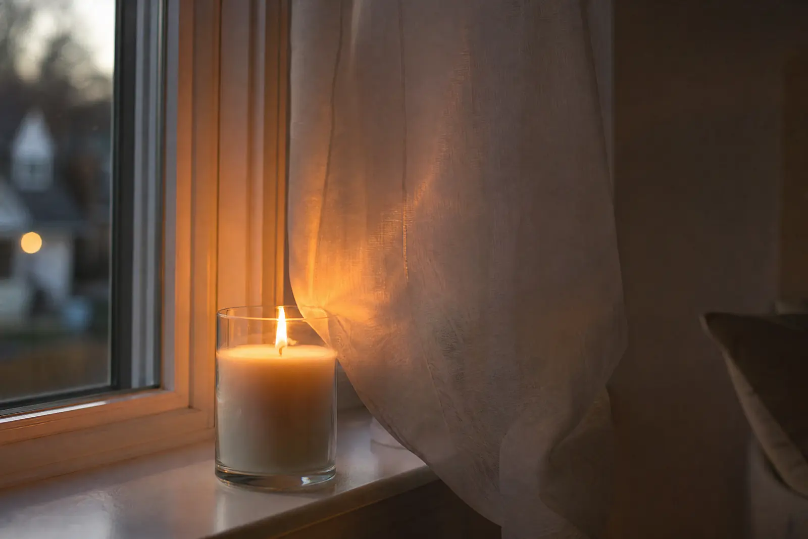 Lit jar candle on a windowsill with a sheer curtain hanging close to the flame